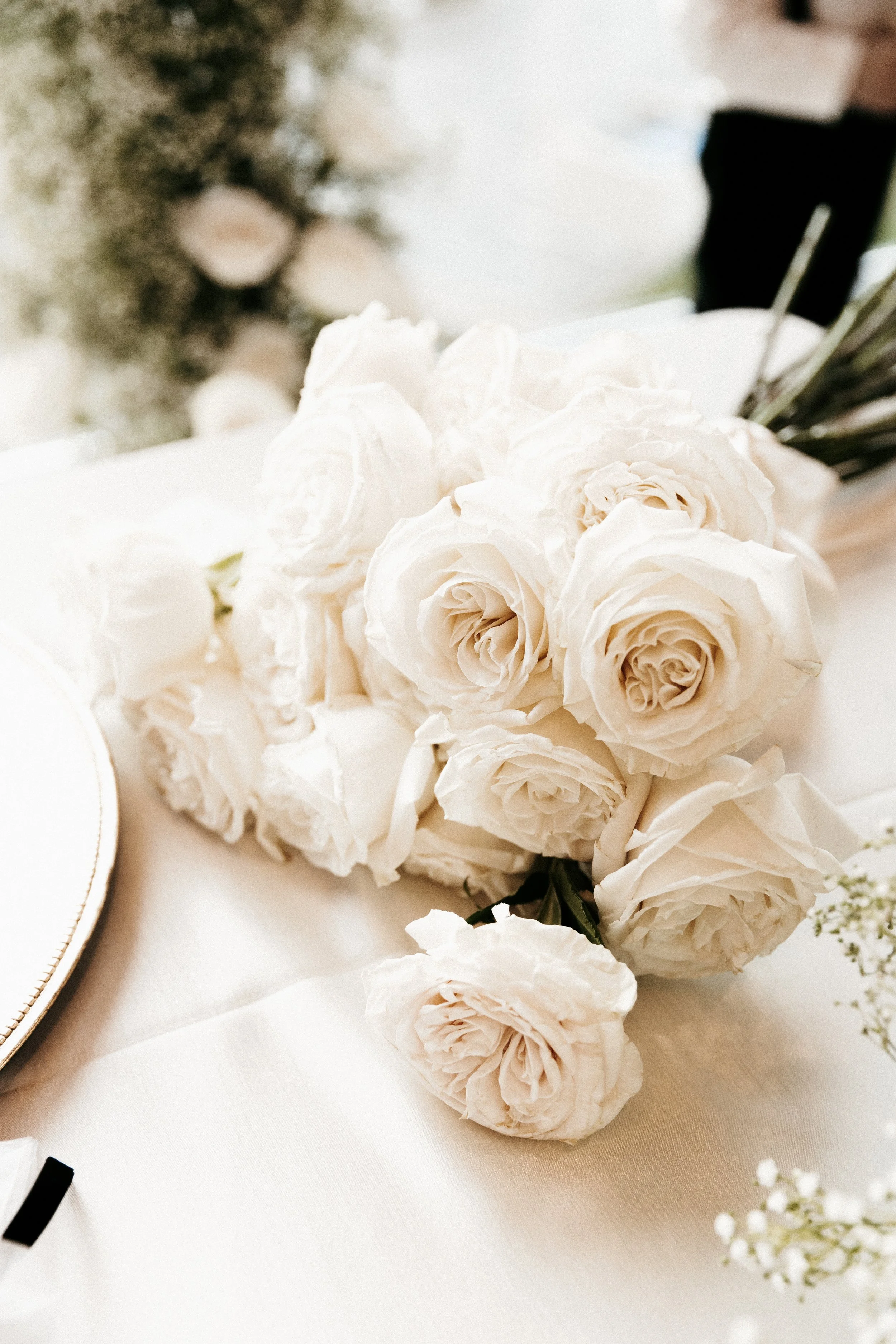 A bouquet of white roses resting on a white tablecloth with a gold-edged plate partially visible on the left.