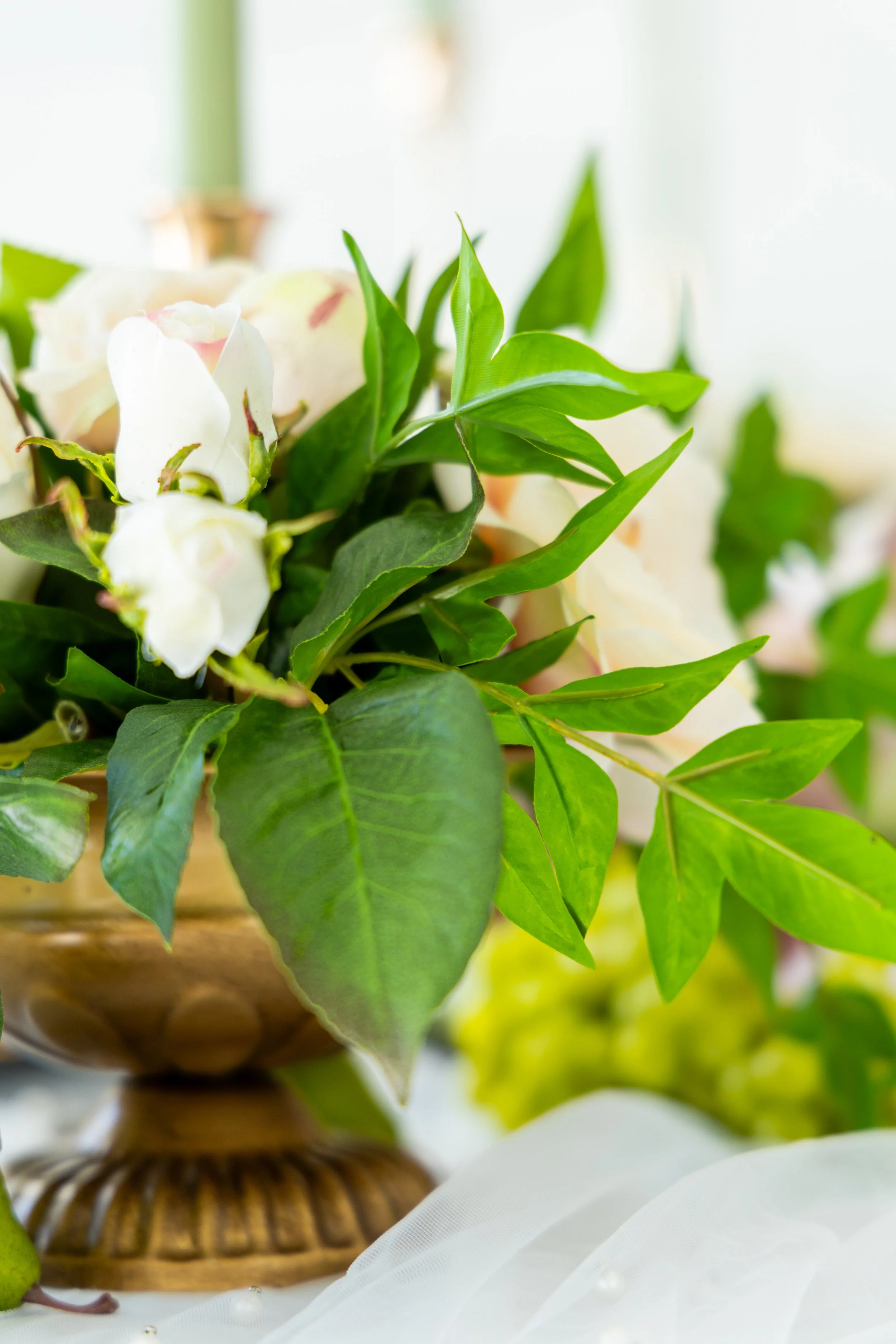 A close-up of a plant with green leaves in a decorative wooden bowl, with white fabric and pearl accents in the background.