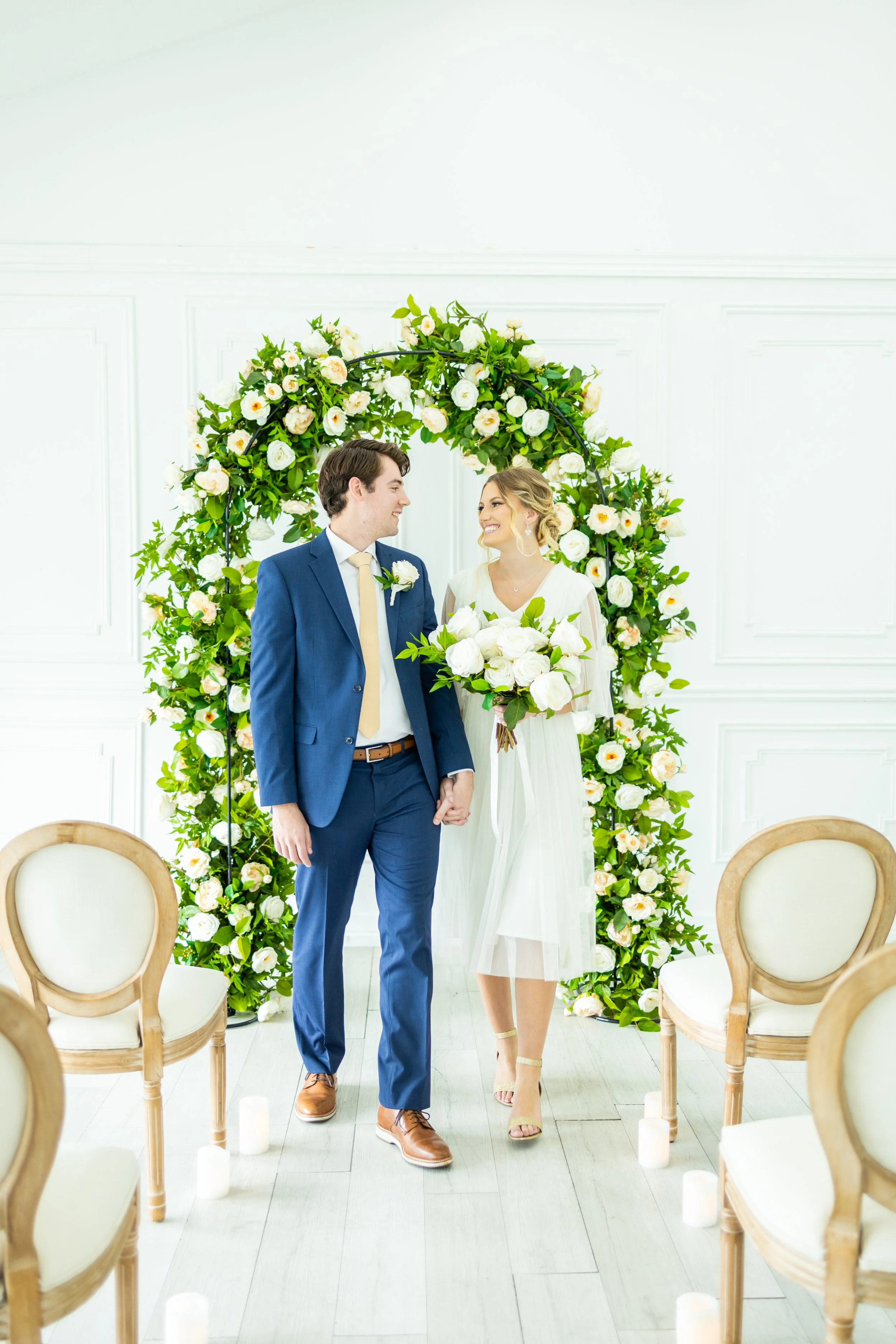 A bride and groom holding hands, standing under a floral wedding arch inside a bright white room, surrounded by empty chairs.