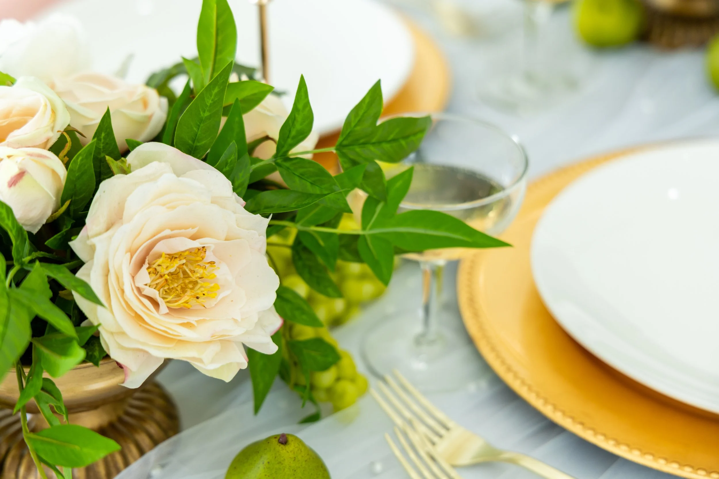 Close-up of a floral arrangement with white and pale pink roses and green leaves, on a table setting with a yellow plate, white dish, wine glass, and a pear in the foreground.