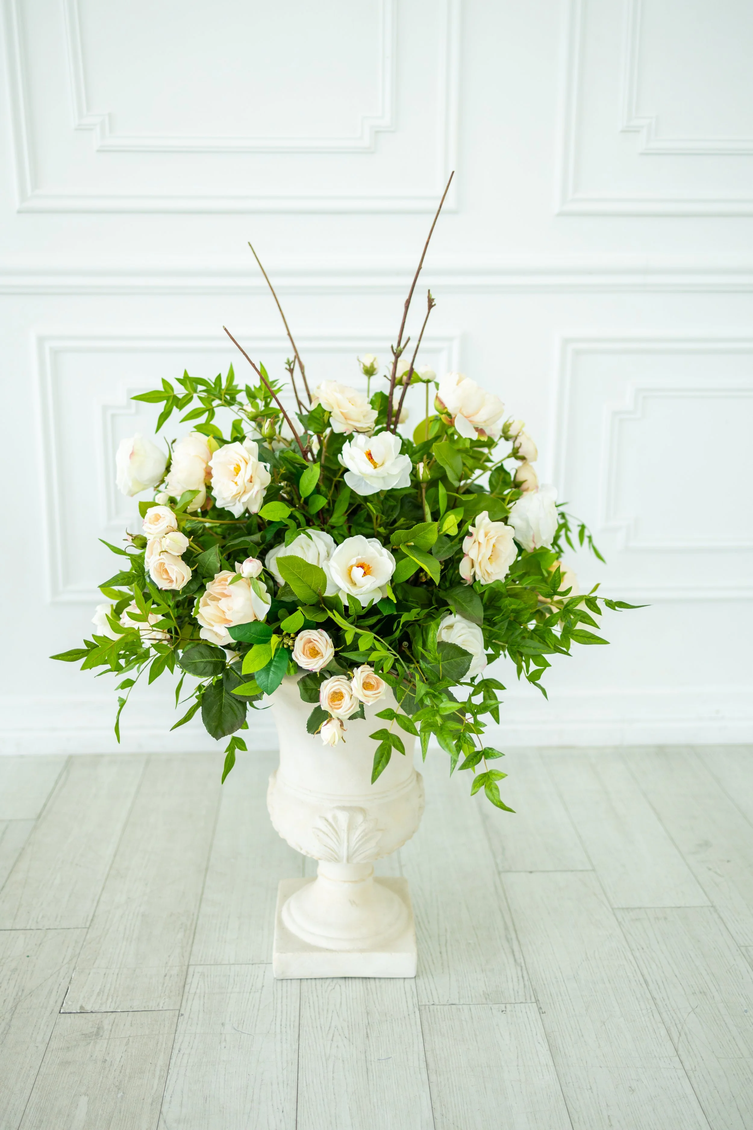 A tall white urn with an ornate base, filled with an arrangement of white and cream-colored flowers with green foliage, set against a white paneled wall and light-colored tiled floor.
