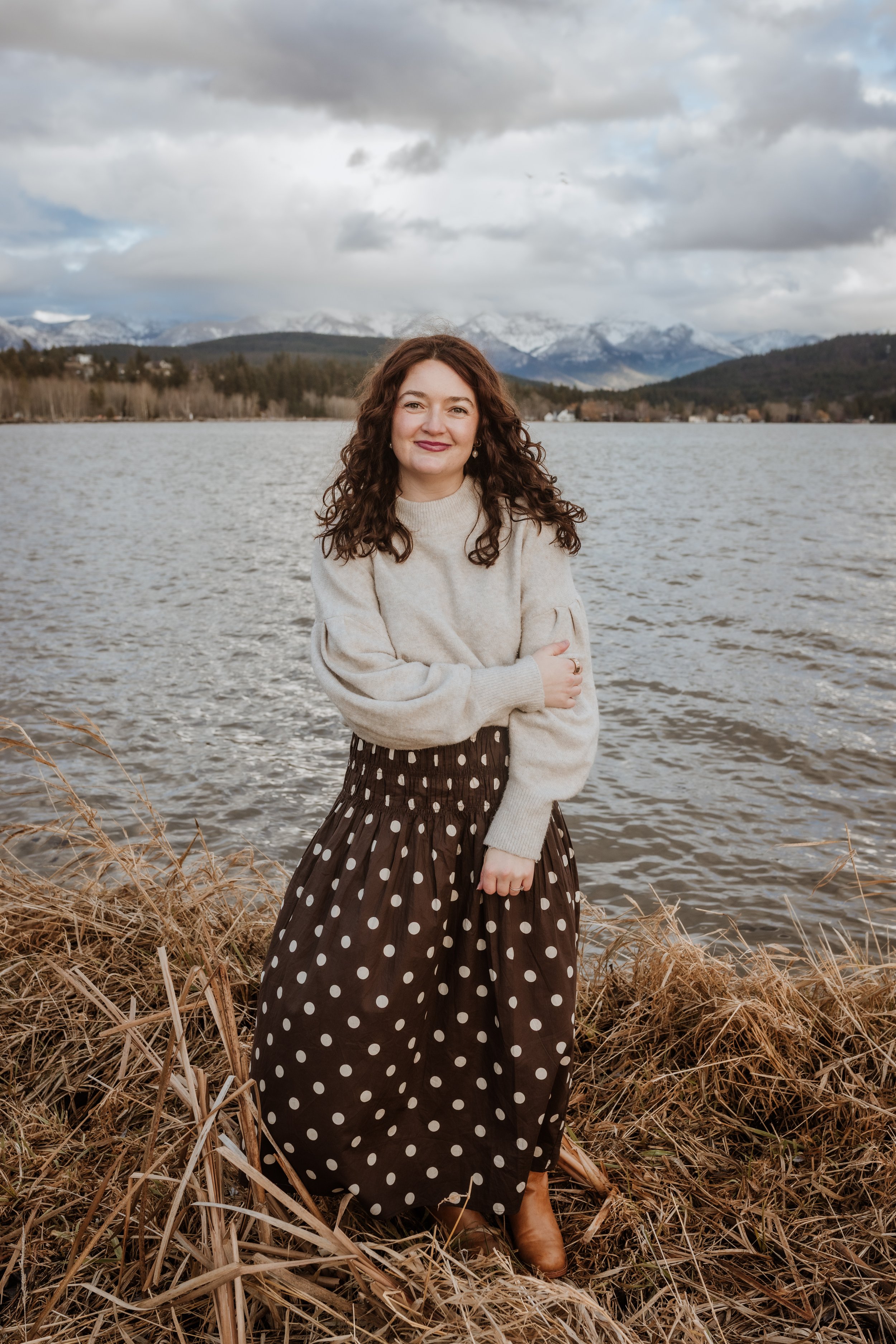 A woman with curly brown hair standing by a lake with mountains in the background. She is wearing a beige sweater, a brown skirt with white polka dots, and tan boots. The scene is outdoors during a cloudy day.