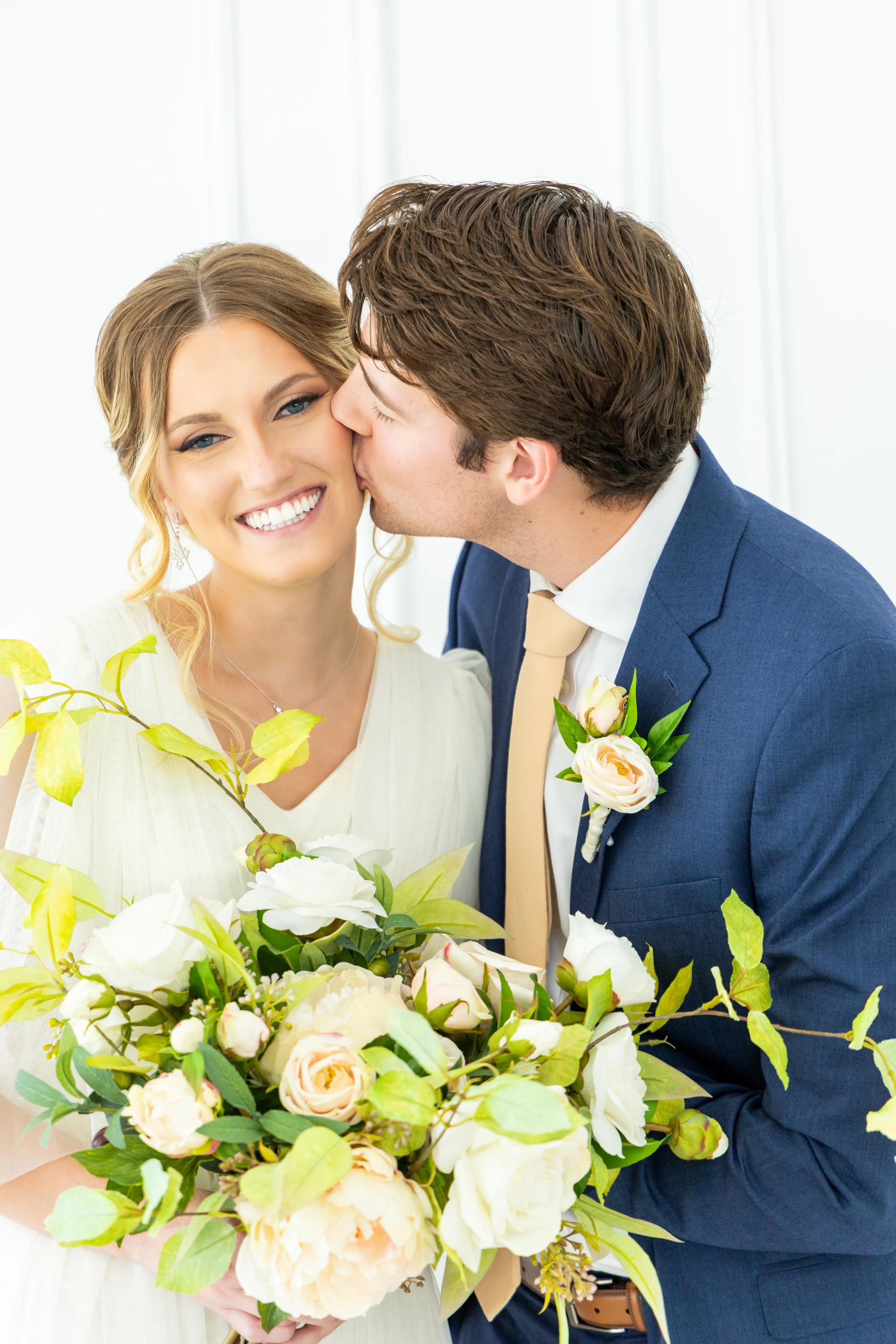 A smiling bride with blonde hair and blue eyes is being kissed on the cheek by a groom with brown hair, both dressed in wedding attire, with a bouquet of white and cream flowers and green foliage in the foreground.