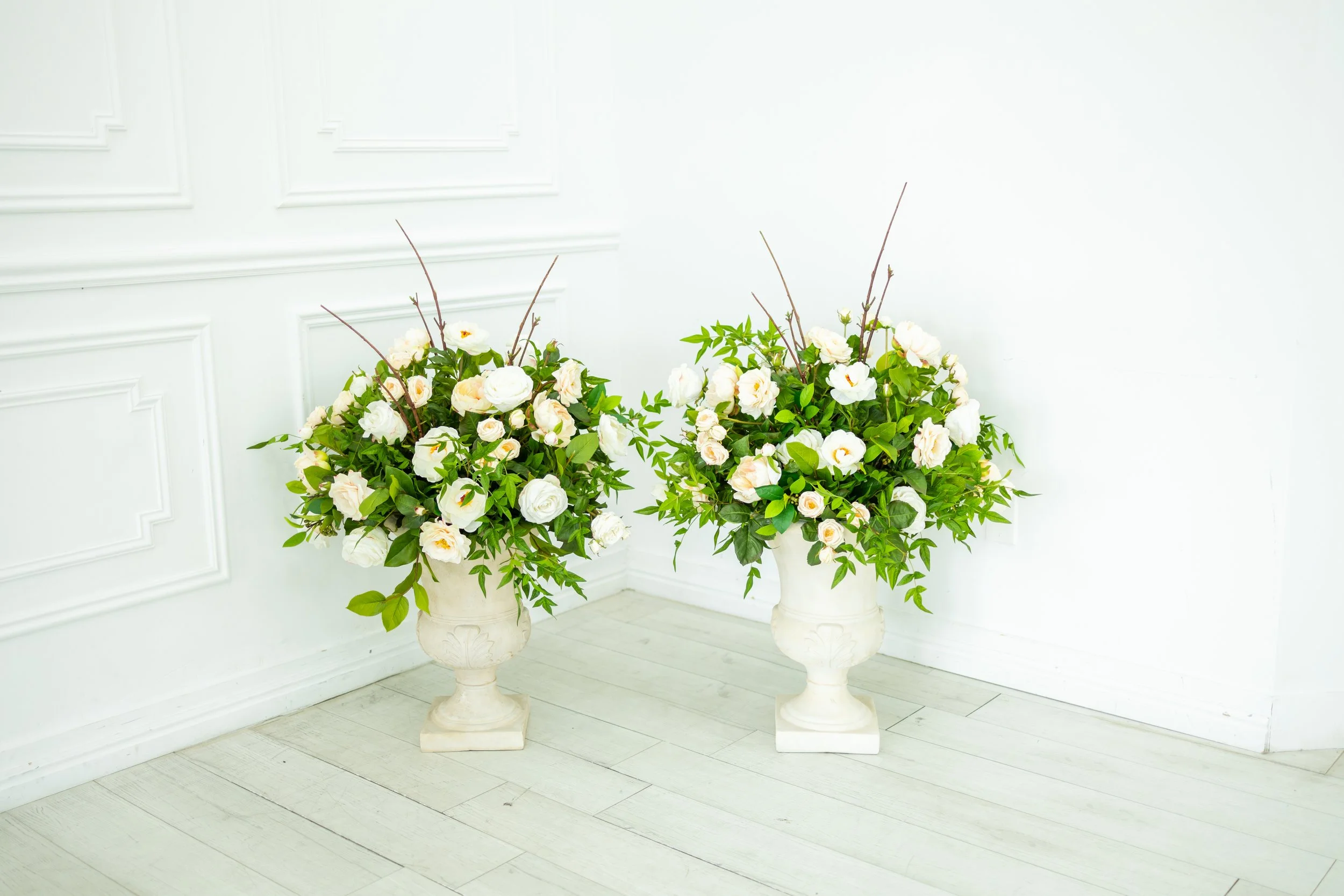 Two elegant white flower arrangements in tall vases placed on the floor in a softly lit room with white walls and wooden flooring.