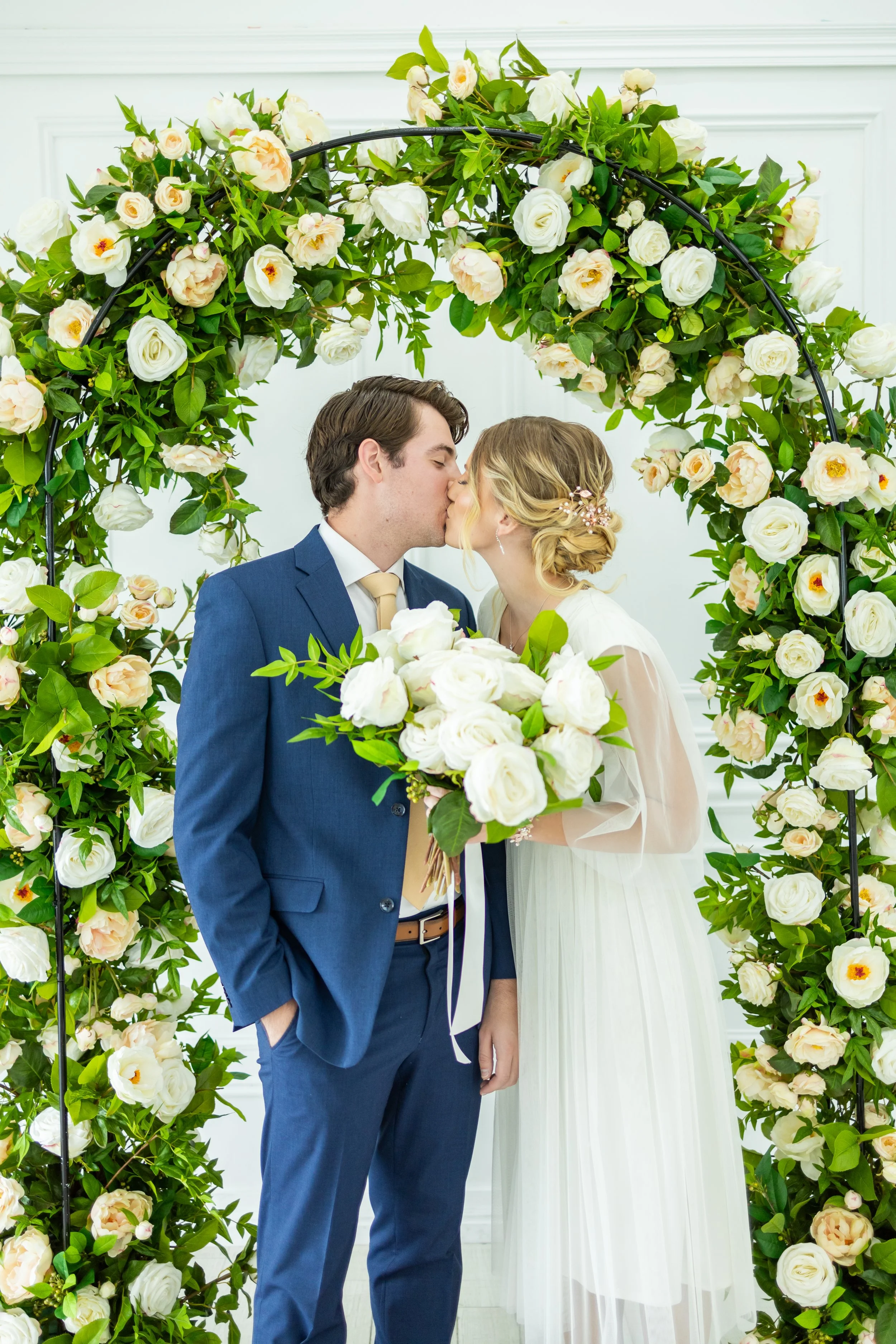 A couple sharing a kiss at their wedding ceremony under a floral arch with white and light pink roses.