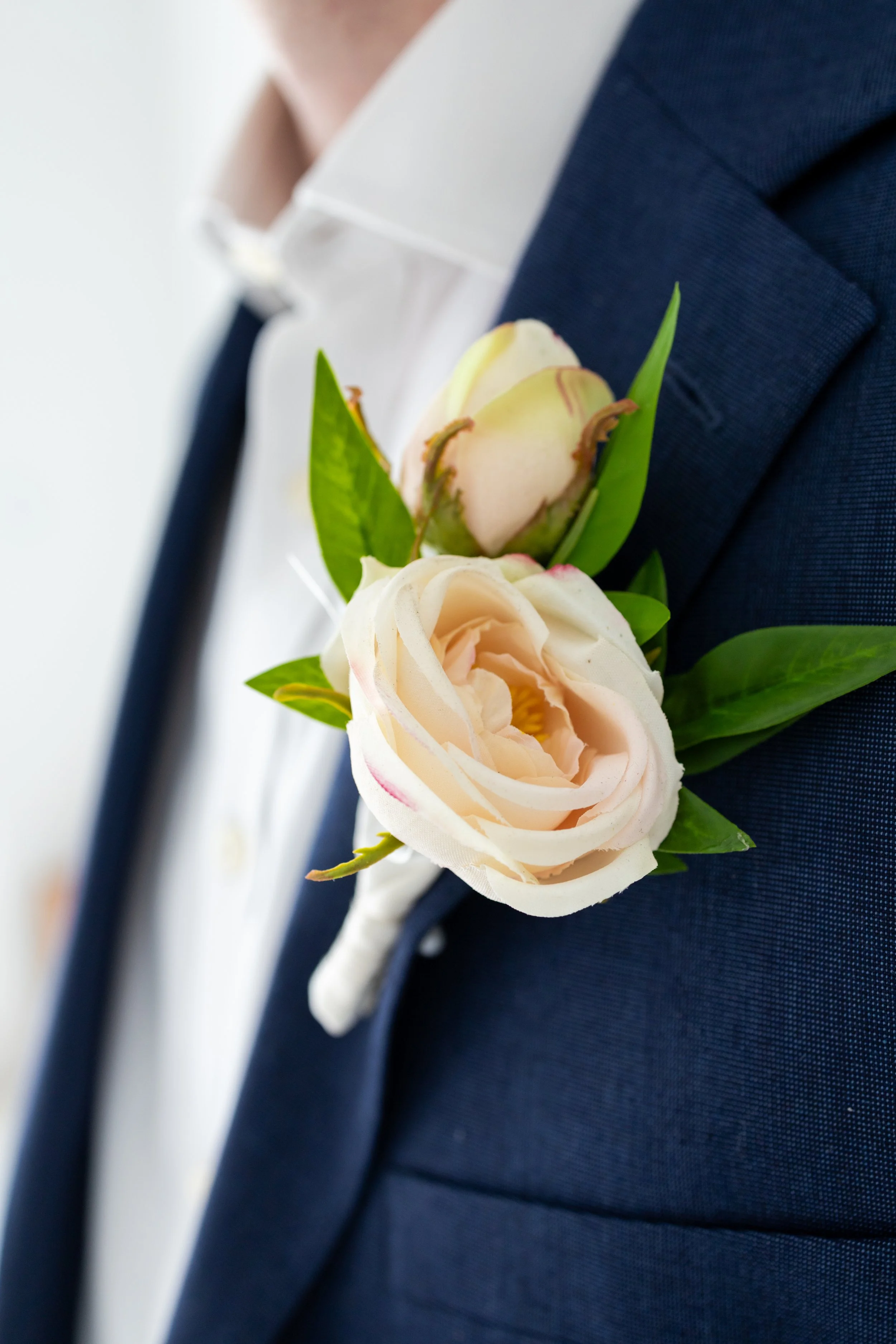 Close-up of a man wearing a dark blue suit with a white shirt and a dark tie, sporting a boutonnière with cream-colored roses and green leaves.