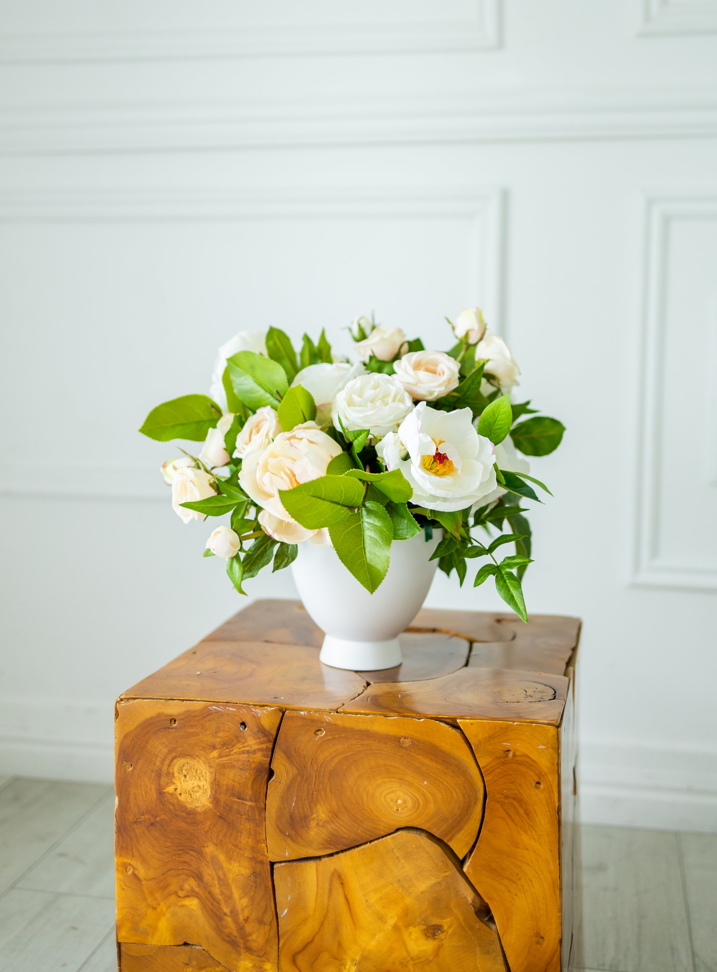 A white ceramic vase filled with white roses and green leaves, placed on a carved wooden table against a white paneled wall background.