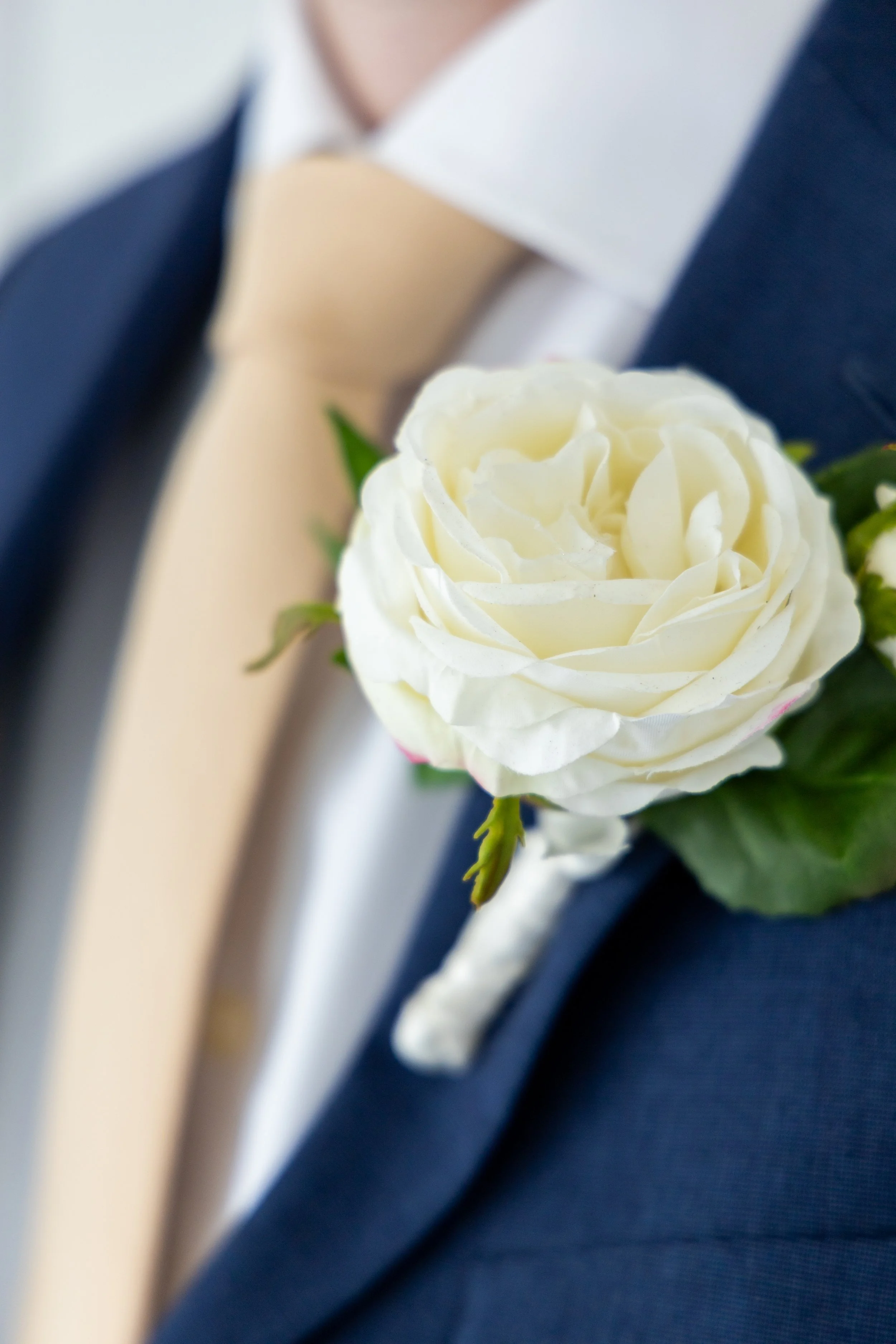 Close-up of a white rose boutonniere worn on a navy blue suit jacket, partially obscured by a beige tie and white dress shirt.