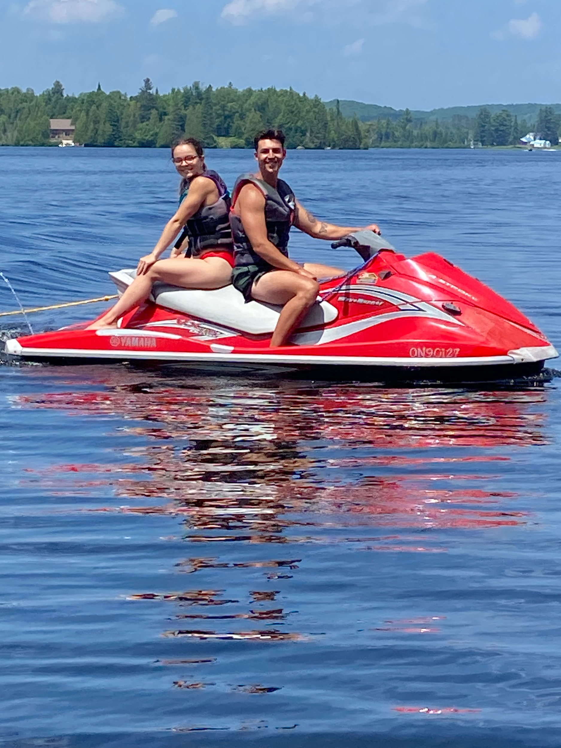 A man wearing sunglasses and a black life vest is standing on a red and white jet ski on a body of water during daytime. He is holding onto the handlebar and smiling. The background shows a tree-lined shoreline and a blue sky.