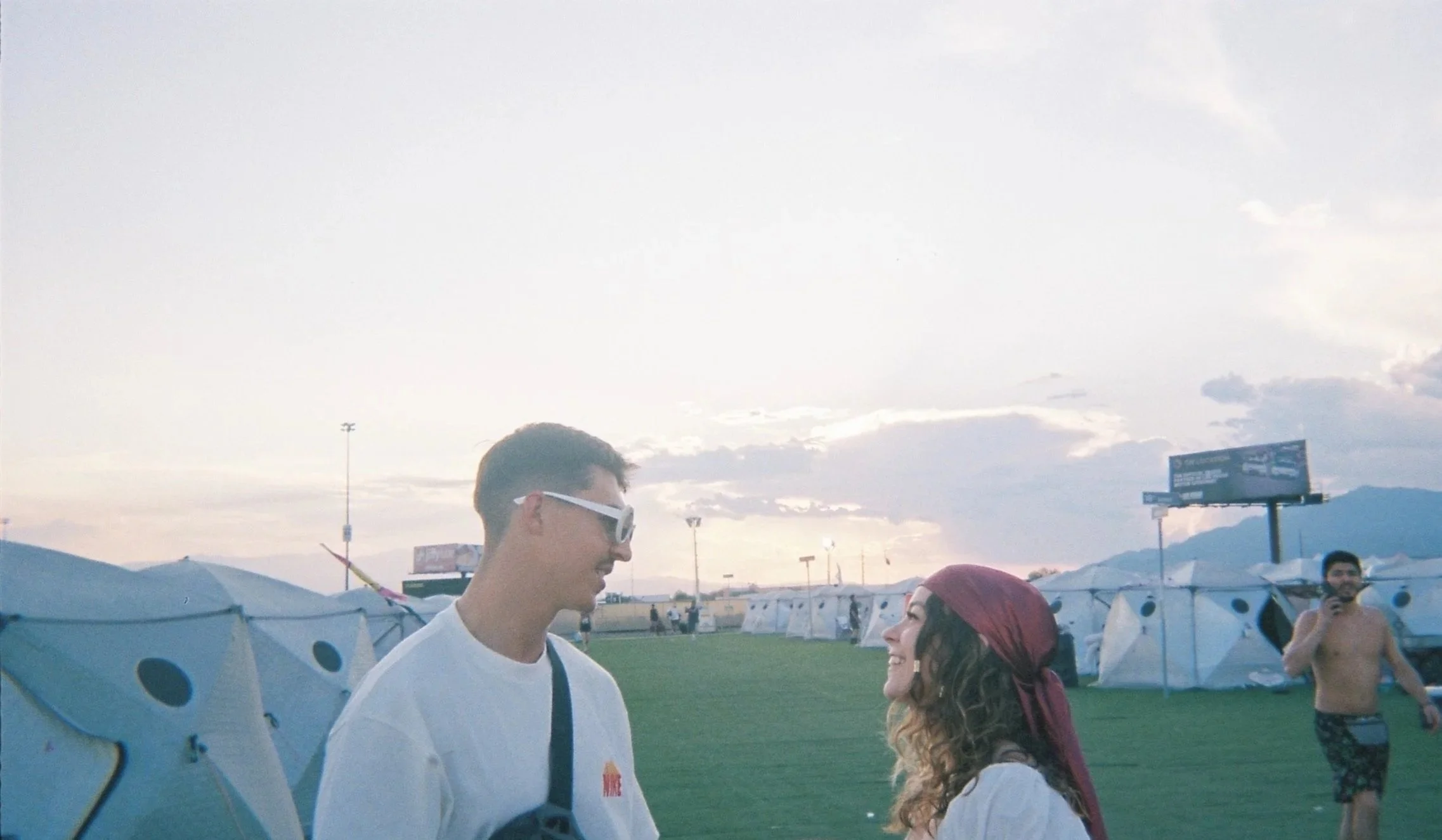 A young man with sunglasses talking to a woman with a red bandana at an outdoor event during the evening. Tents are visible in the background.