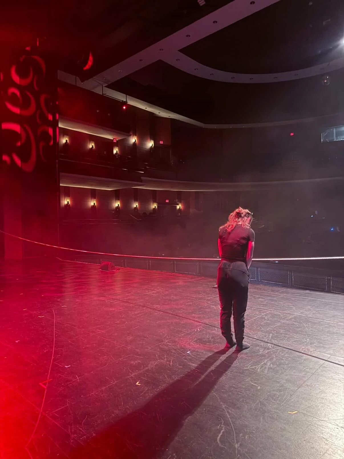 A woman stands on a stage in a theater, facing away from the camera, with empty seats and balconies in the background, illuminated by red and warm lights.