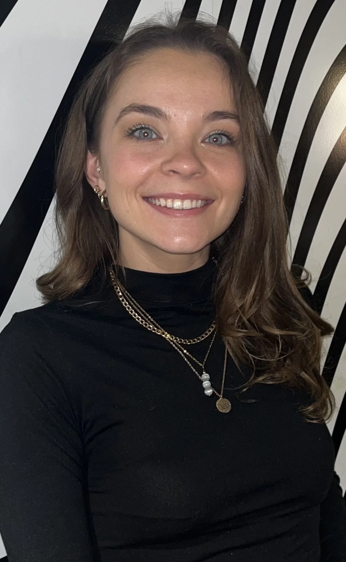 A young woman with brown hair and blue eyes smiling, wearing a black turtleneck sweater and layered gold necklaces, standing in front of a black and white striped background.