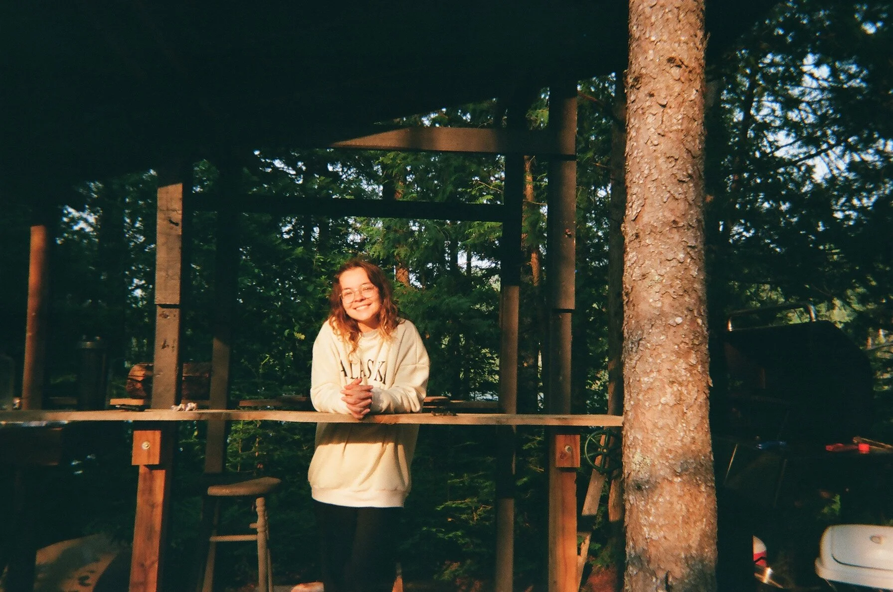 A young woman with curly hair and glasses smiling, standing outside on a wooden deck surrounded by tall trees and greenery.