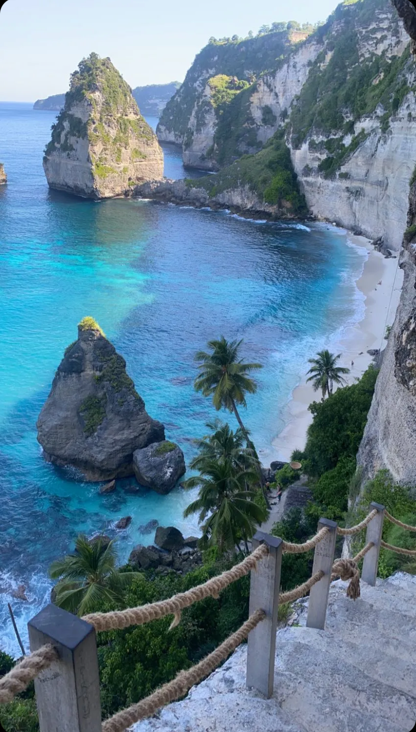 Coastal scene with cliffs, clear turquoise water, large rock formations, and palm trees along the shoreline.