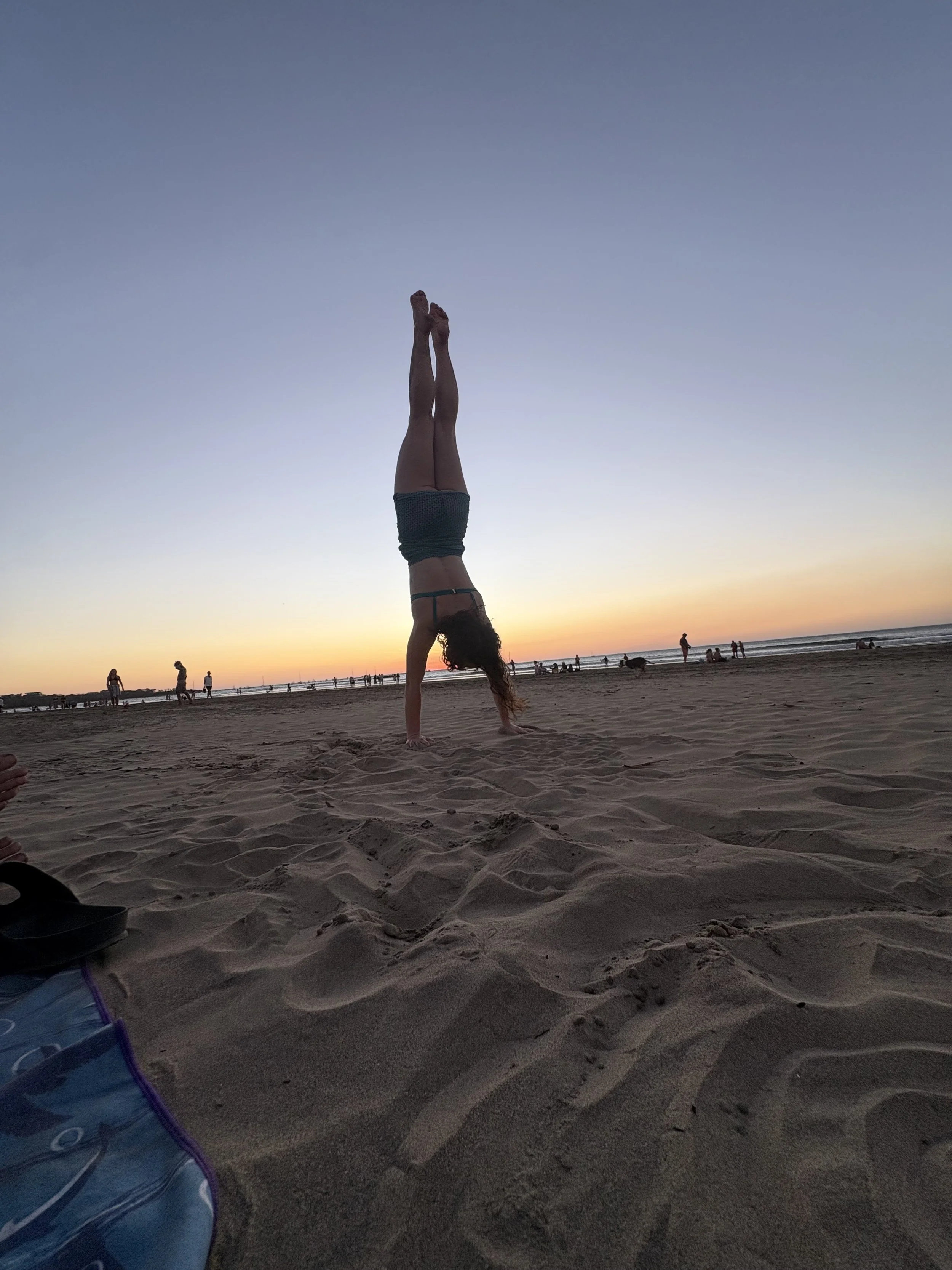 A person doing a handstand on the beach at sunset, with various people and dogs in the background, and a beach bag in the foreground.