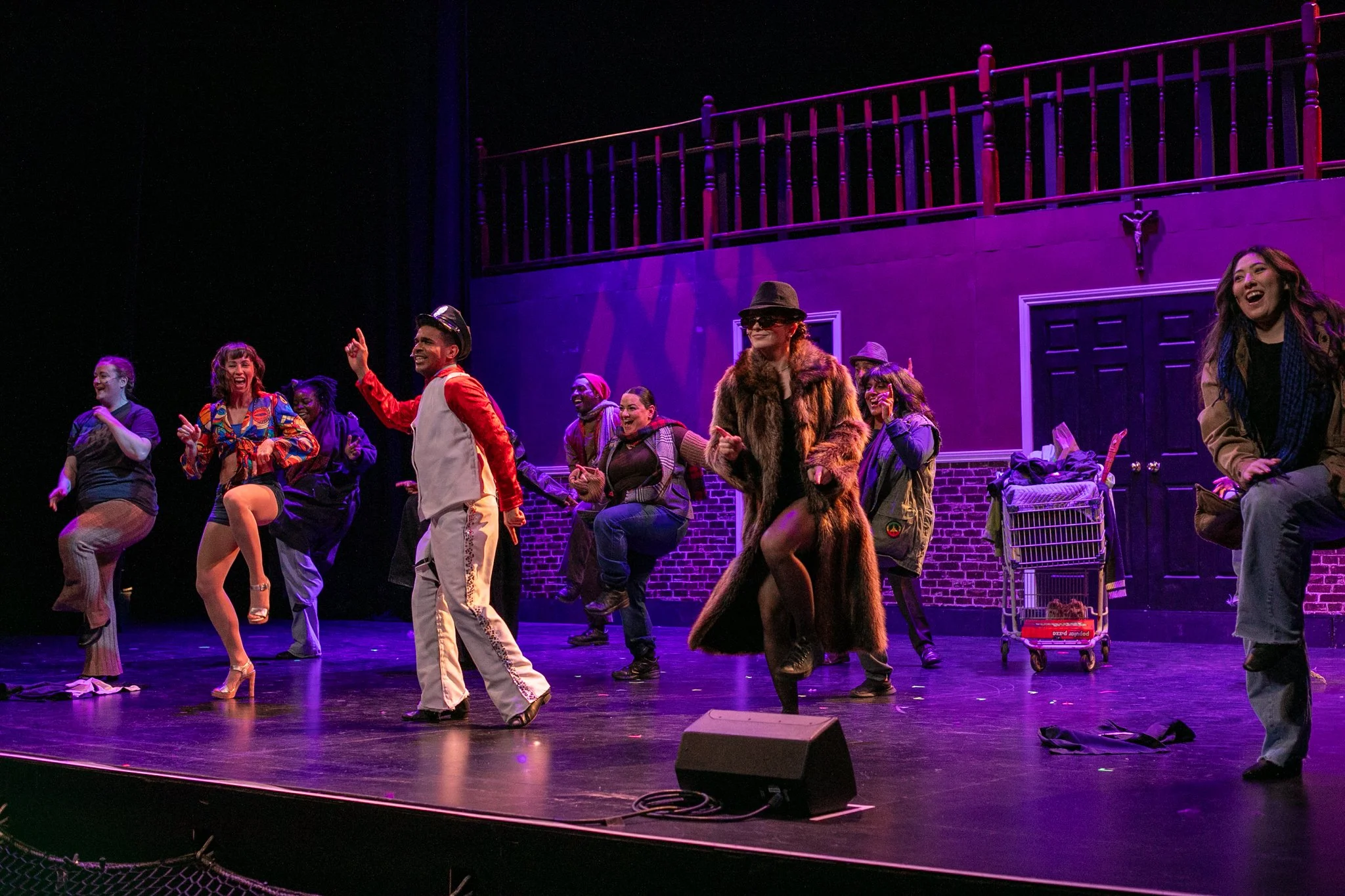 A theatrical stage with diverse performers dancing and smiling, dressed in colorful and eccentric costumes, under purple stage lighting, with a set resembling a neighborhood scene with a building and balcony in the background.