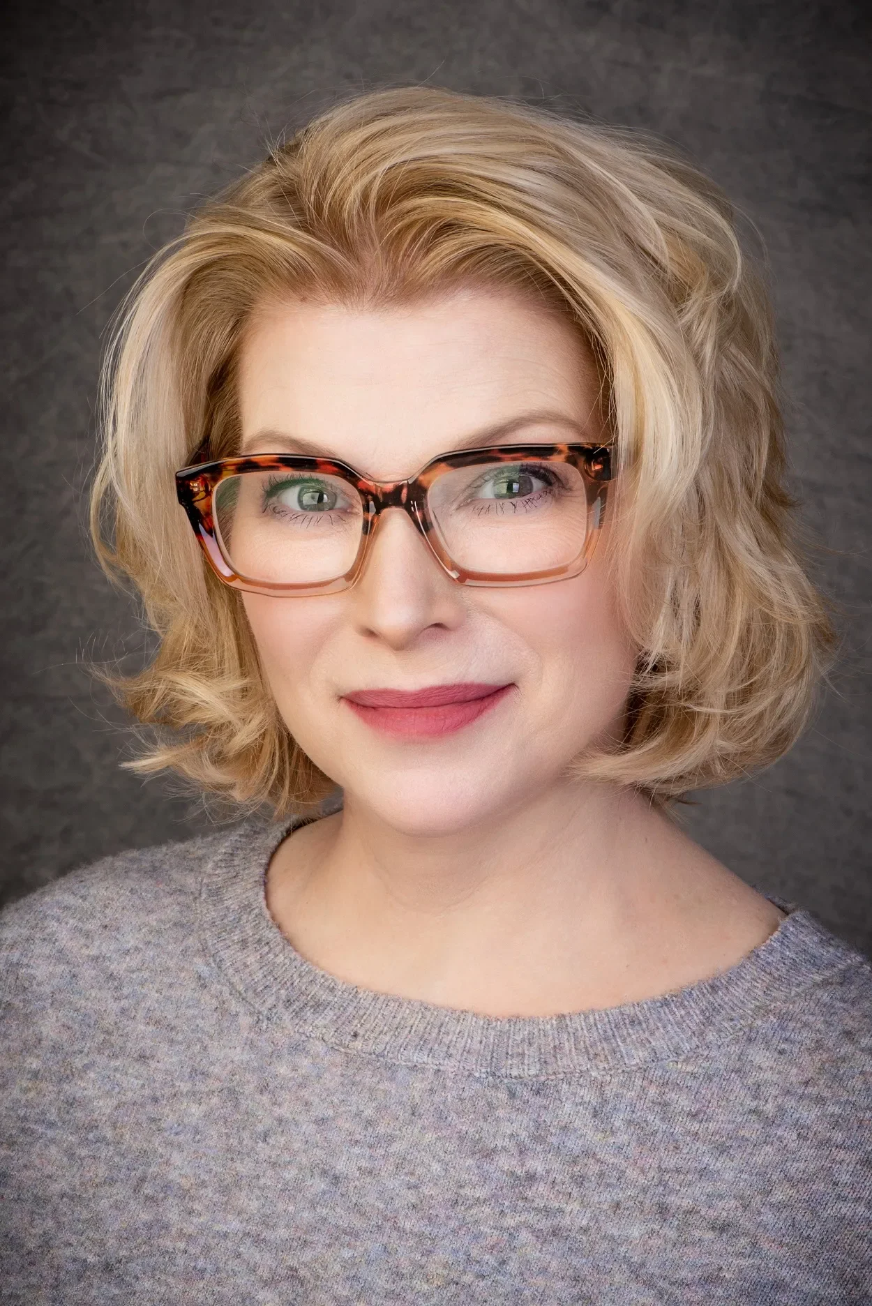 A woman with blonde, wavy hair wearing tortoiseshell glasses and a gray sweater, smiling softly against a gray background.