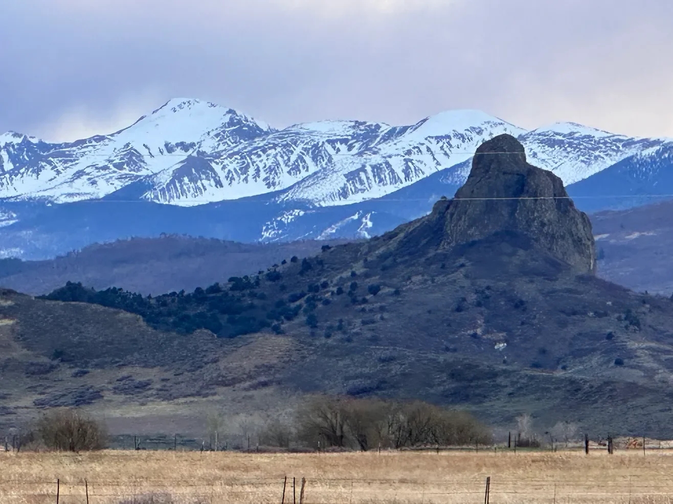 Scenic landscape with a large rock formation in the foreground, rolling hills, and snow-capped mountains in the background under a cloudy sky.