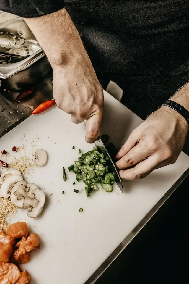 Person chopping green onions on a white cutting board, with sliced mushrooms, small orange vegetables, and spices nearby.
