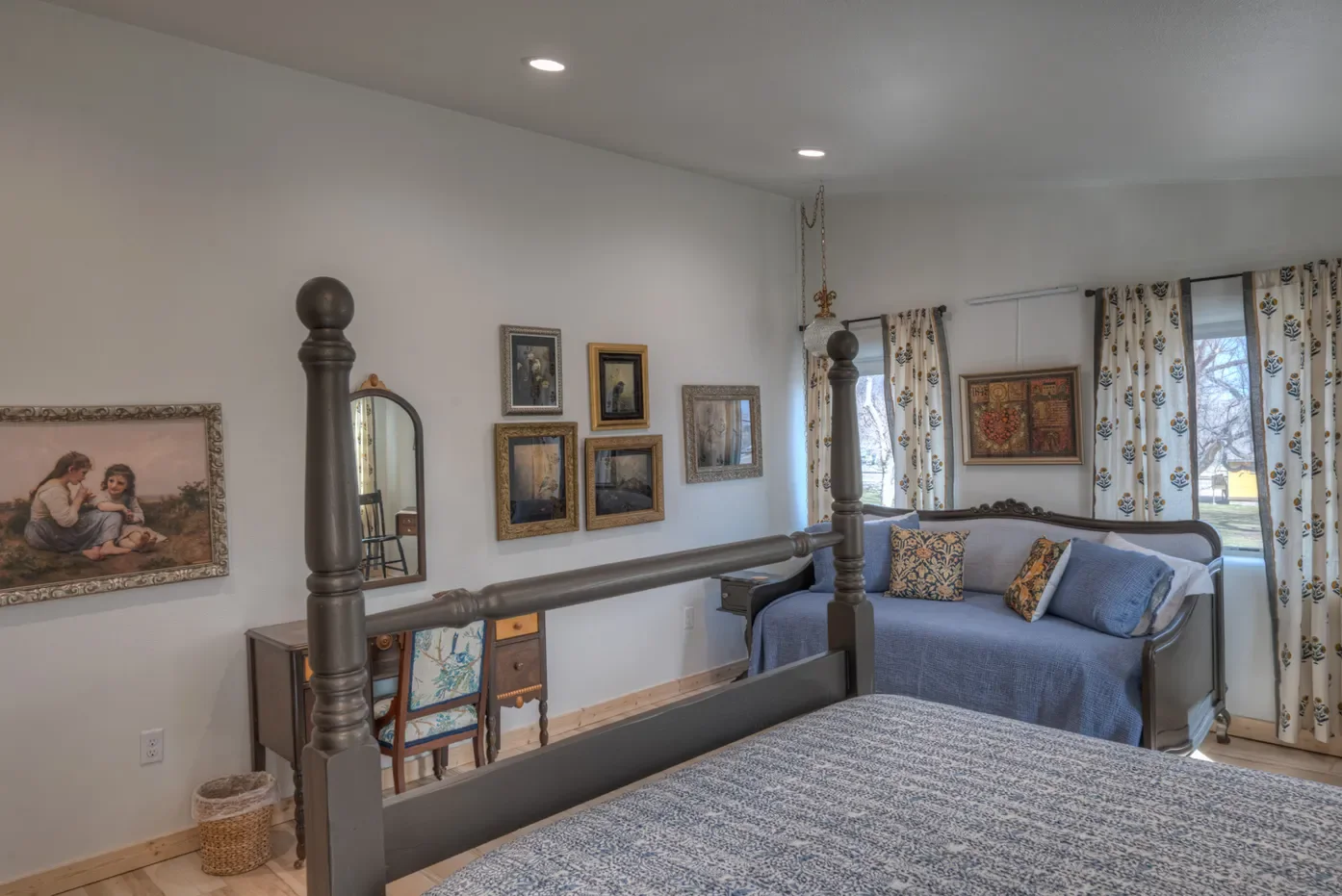 A bedroom with a gray wooden bed and a gray sofa with decorative pillows, framed artwork on the walls, and a window with floral curtains.