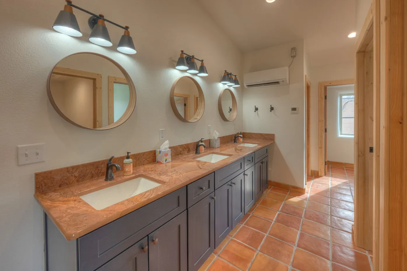 A spacious bathroom with three sinks on a brown marble countertop, three round mirrors above each sink, and overhead lighting fixtures. The room has a terracotta-tiled floor, light-colored walls, and a wooden accent wall.