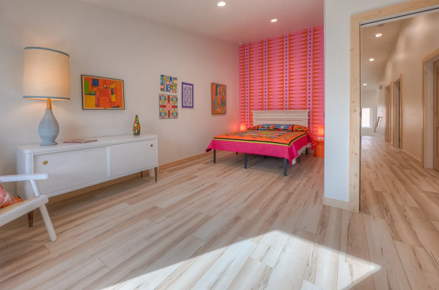 A bedroom with a white bed, pink and orange geometric patterned accent wall, and colorful artwork on white walls. There is a white dresser with a lamp and a small decorative bottle, and a wooden chair with a cushion. The room has light-colored wood flooring and recessed lighting.