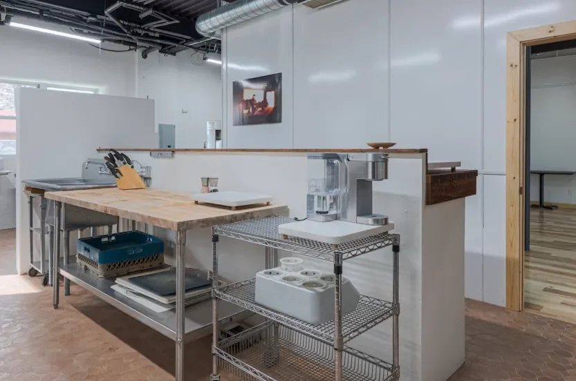A kitchen workspace with a wooden countertop, metal shelves with various kitchen items, a water filter, and a knife block, in an industrial-style space with white walls and exposed ductwork.