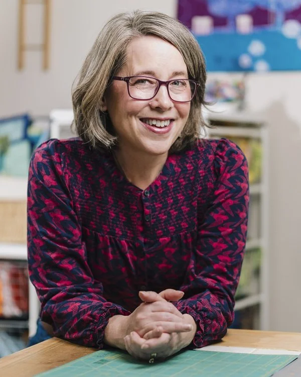 A smiling woman with glasses, wearing a red and navy patterned blouse, sitting at a table in a colorful classroom or craft room.