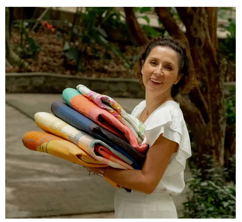 Woman smiling and holding a stack of colorful folded blankets outdoors near trees.