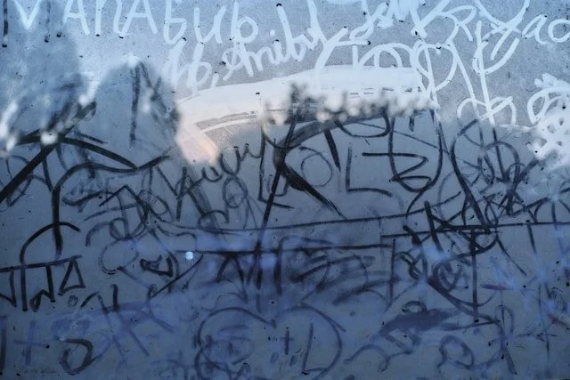 A glass window covered with various overlapping black and white graffiti and scribbles, with a reflection of a car and a faint sky visible in the background.
