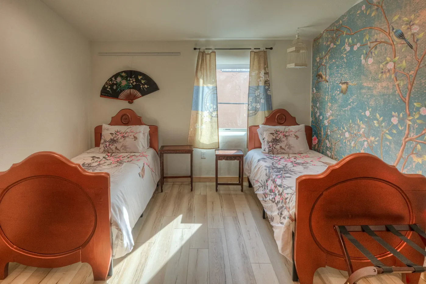Two single beds with floral bedding and wooden headboards in a bedroom, with a window between them, floral curtains, a decorative wall fan, and a large floral wallpaper mural of birds and cherry blossoms.