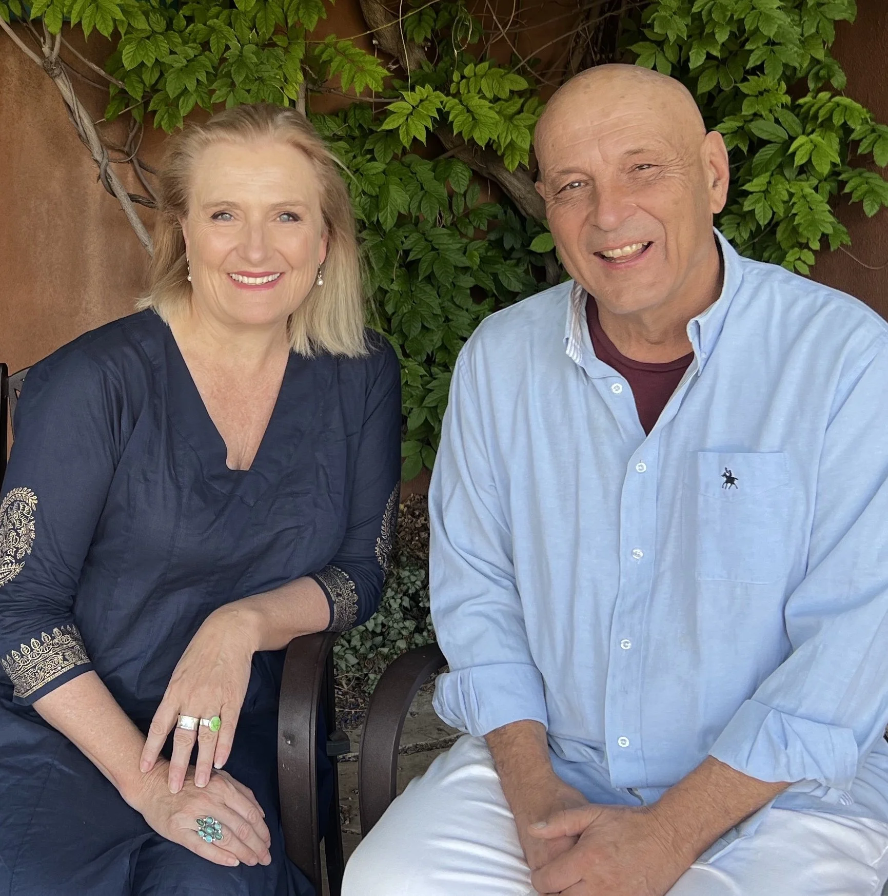 A smiling middle-aged woman with blonde hair wearing a navy blue blouse with embroidery on the sleeves, sitting outdoors next to an older bald man in a light blue button-up shirt, in front of green leafy plants and a brown wall.