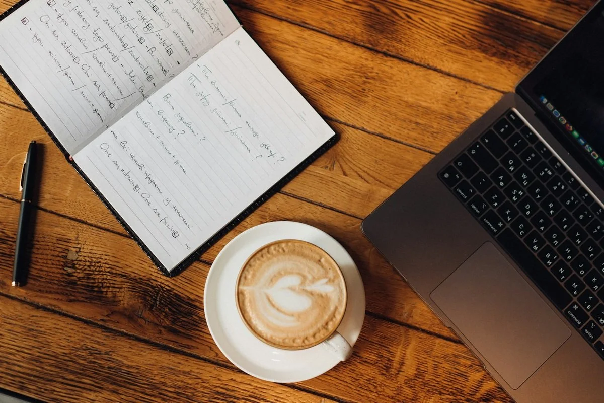 Open notebook with handwritten notes, black pen, white coffee cup with latte art, and part of a black laptop on a wooden table.