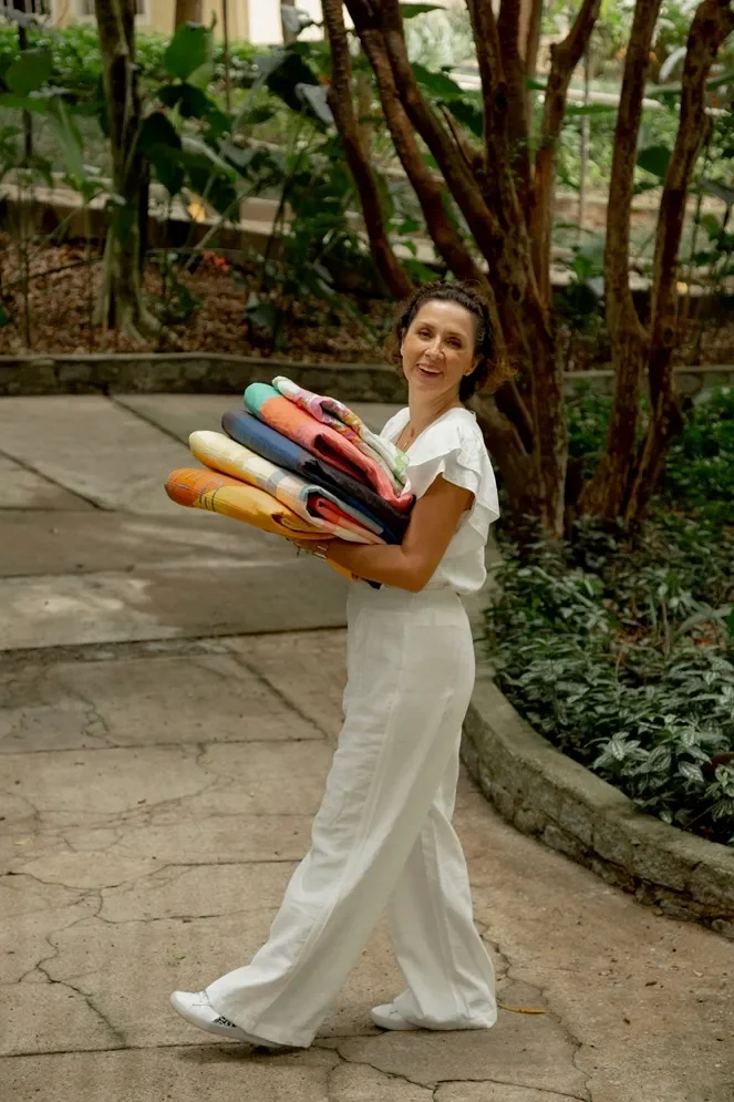 A woman smiling and holding a large stack of colorful folded towels or blankets, walking on a paved path in a lush indoor botanical garden.