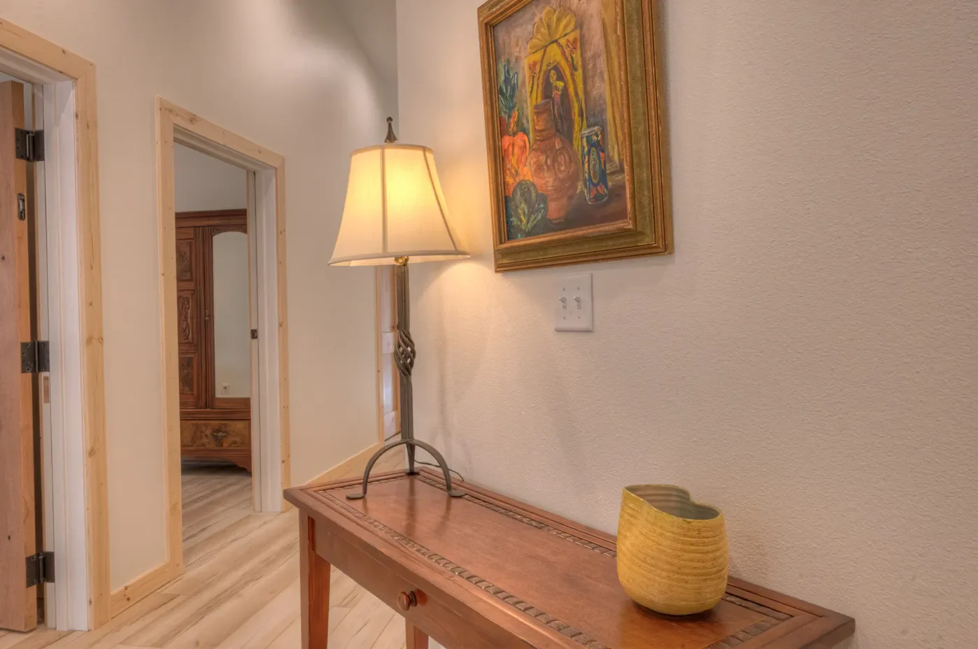 A hallway with cream-colored walls, featuring a side table with a yellow ceramic vase, a tall lamp with a beige shade, and a framed colorful painting of traditional still life. Doorways and wooden trim are visible.