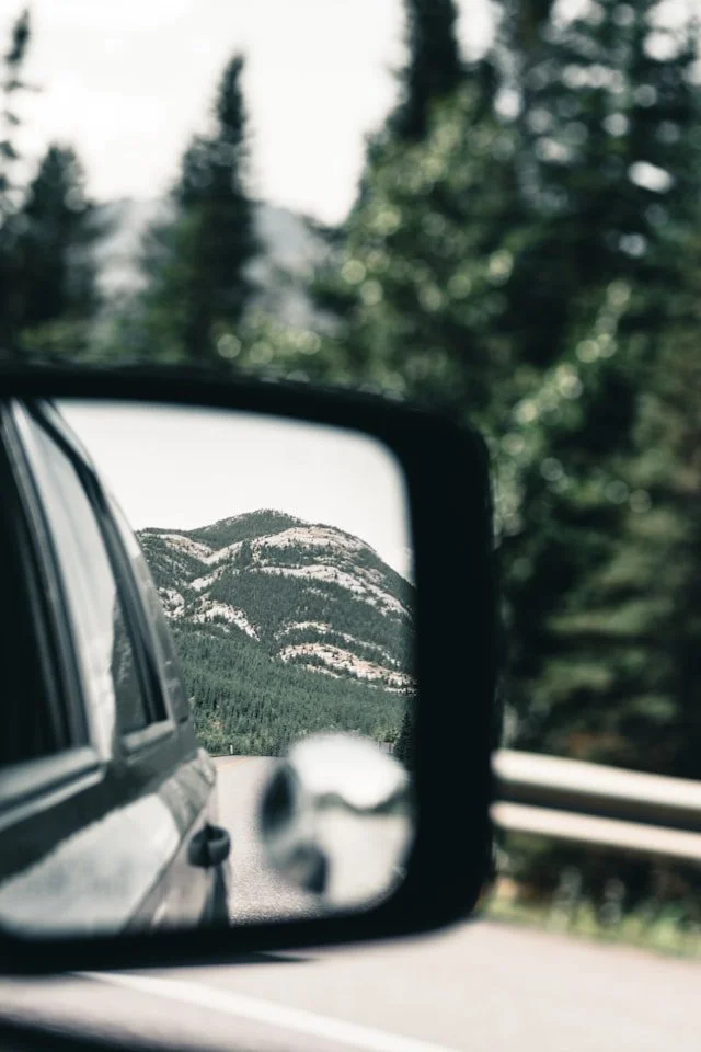 View of a mountain range seen in a car's side mirror, with a portion of the car and green forested hillside visible.