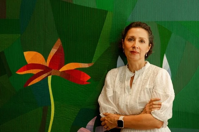 A woman with short dark hair wearing a white blouse, standing with her arms crossed in front of a green mural with a large red and orange flower design.