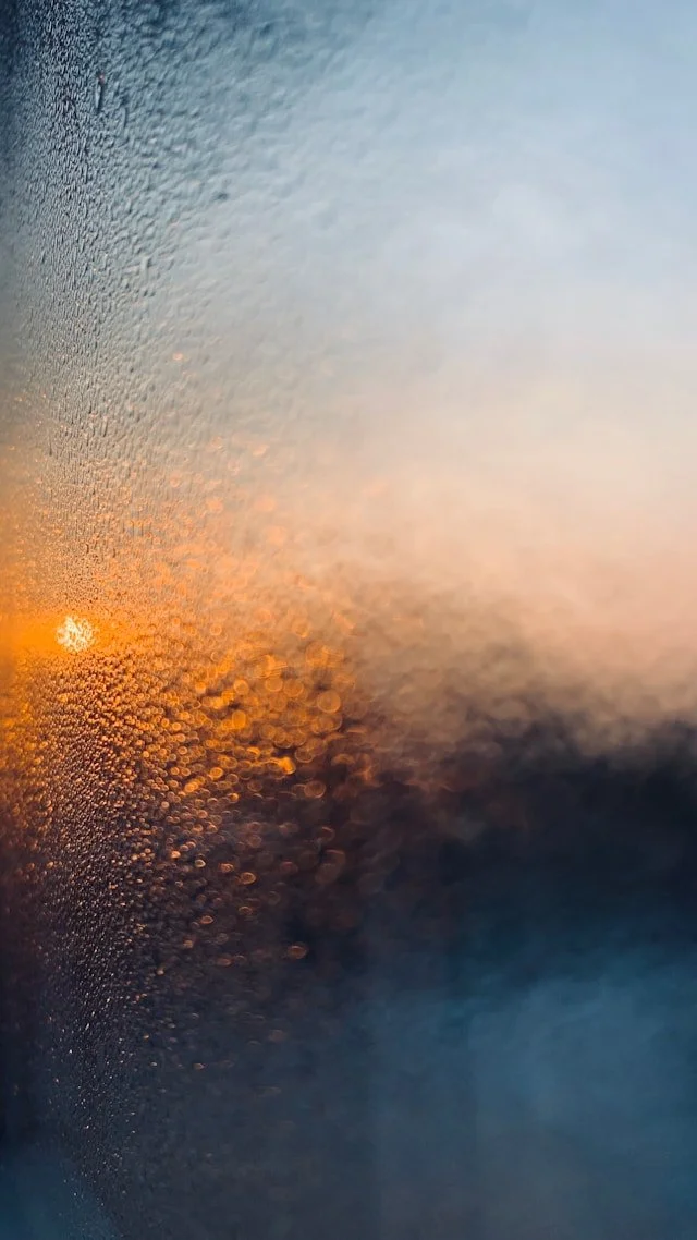 A view through a window with condensation and water droplets, showing a sunset or sunrise with warm orange and yellow hues.