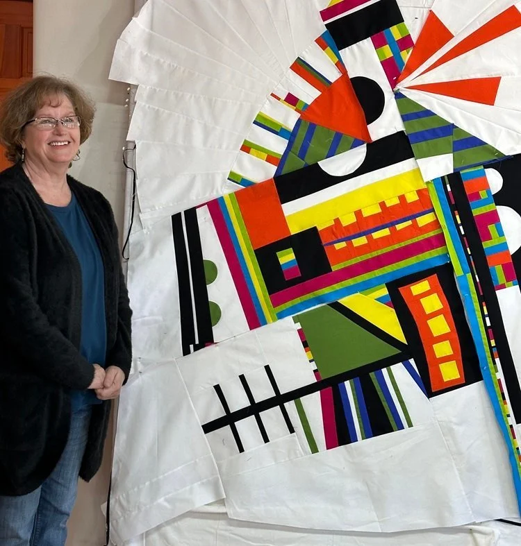 A woman with glasses and curly hair stands beside a colorful abstract geometric quilt with various shapes and patterns, mounted on a white background.