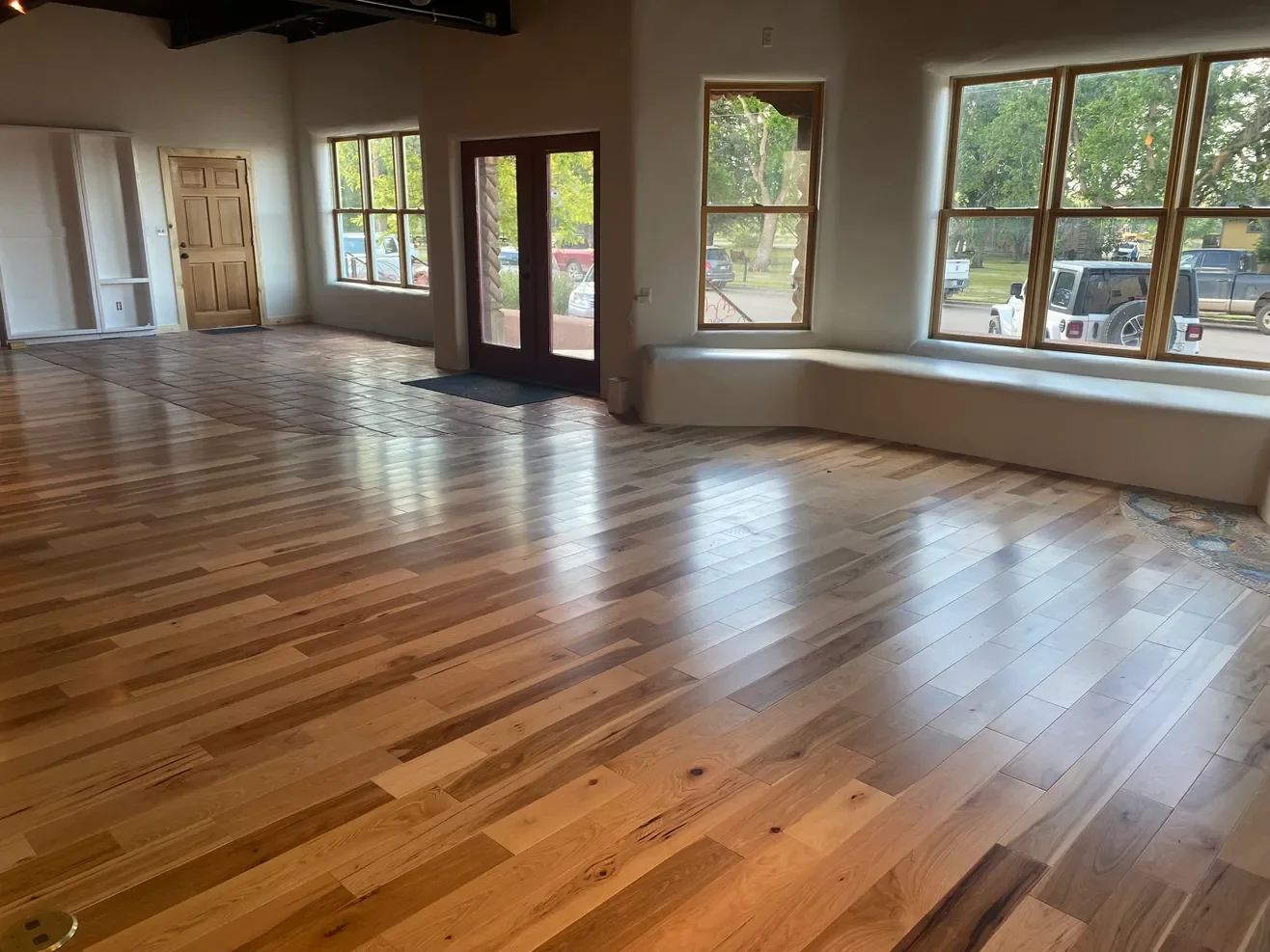 Empty interior space with hardwood floors, large windows, a wooden door, and glass double doors leading outside, with sunlight coming through the windows.