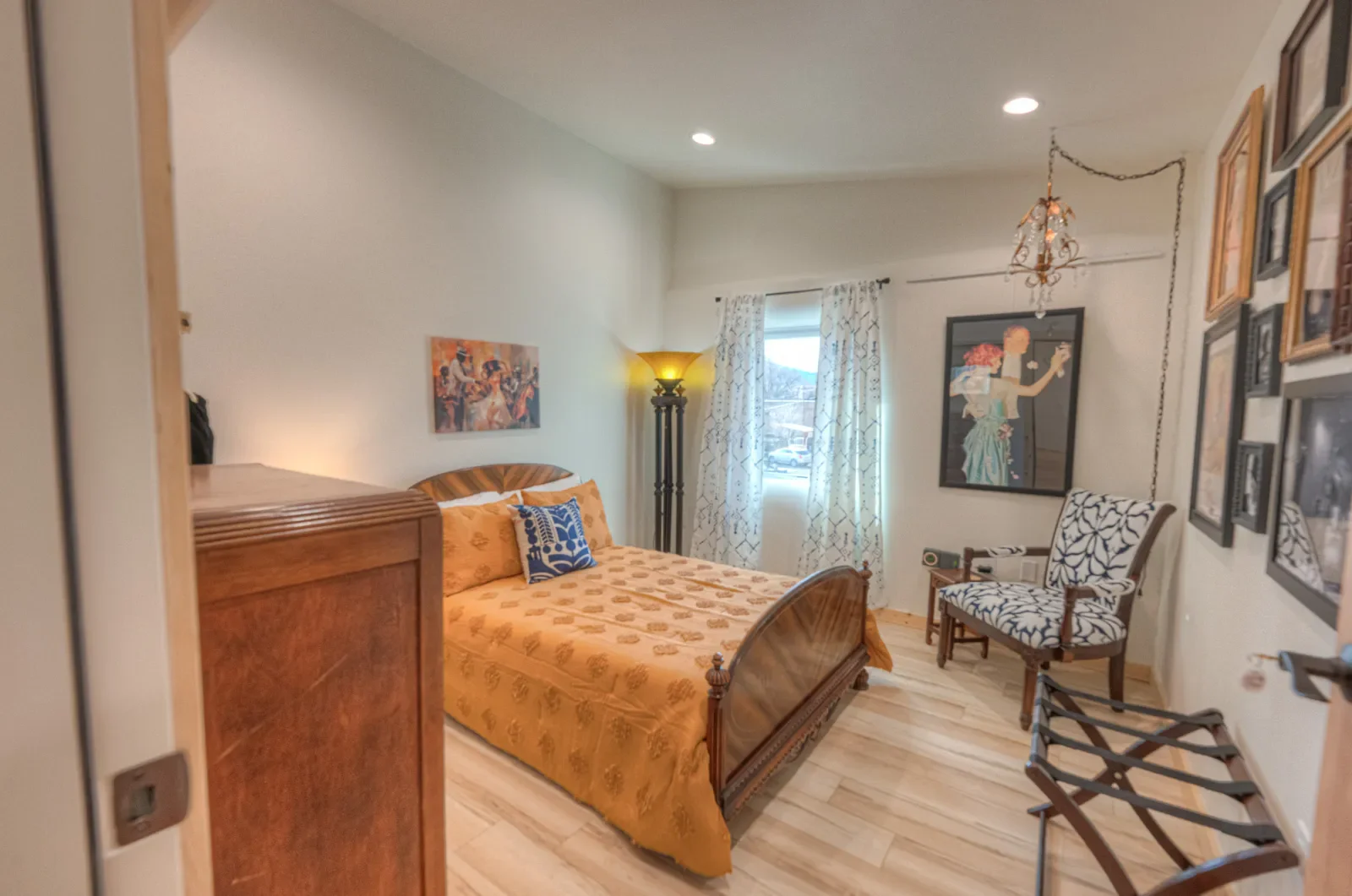 A cozy bedroom with a wooden bed, orange bedding, and patterned throw pillow. There is a window with white curtains, a standing lamp, framed artwork on the walls, and a zigzag-patterned armchair near a small side table.