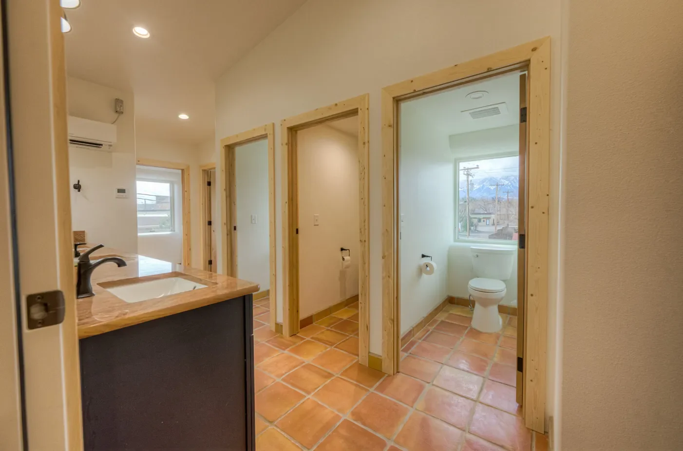 Interior view of a house bathroom with two separate toilet stalls, wood-framed doors, a window showing an outdoor neighborhood scene, terracotta tile flooring, and a partial view of a kitchen with a countertop and sink.