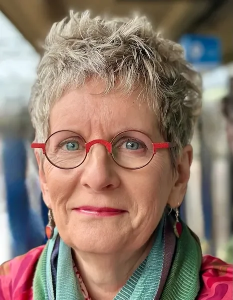 Close-up of an older woman with short, gray hair, wearing red glasses, earrings, and a colorful scarf, smiling near a train window.