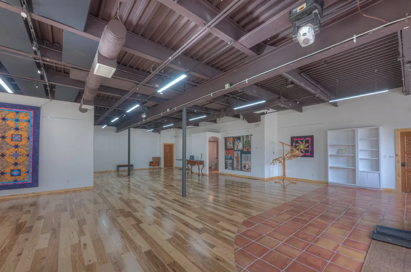 Empty art gallery with hardwood and tile flooring, white walls, colorful quilt on the left, quilted artwork on the right, a coat rack, and ceiling with exposed ductwork and lighting fixtures.