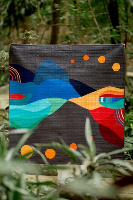 Abstract colorful quilt with geometric shapes and patterns, hanging outdoors among green foliage.