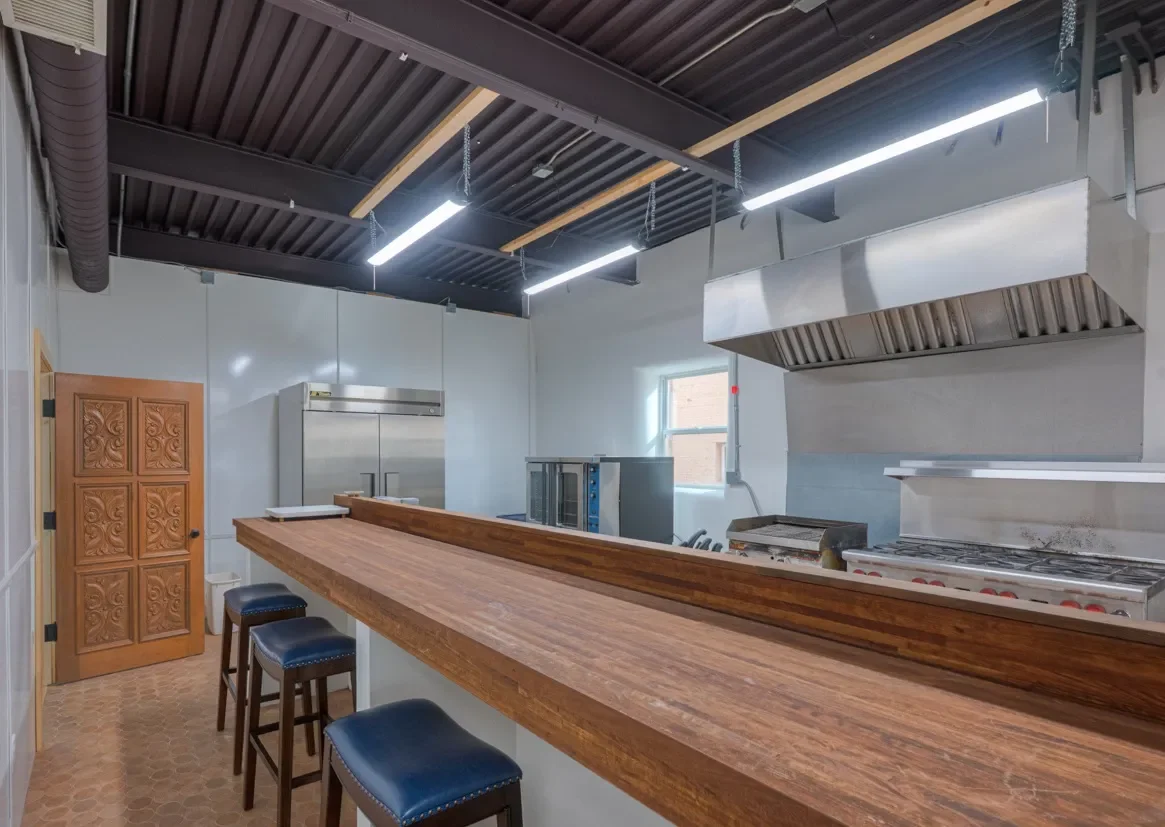 Interior of a commercial kitchen with a wooden bar counter, black stools with blue upholstery, stainless steel appliances, a window, and a decorative wooden door.