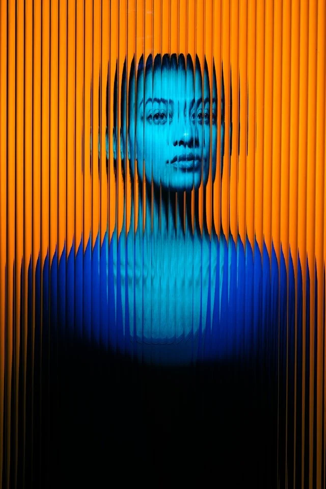 A portrait of a woman, rendered with a blue tint, seen through vertical orange and black slats creating a distorted, layered effect.