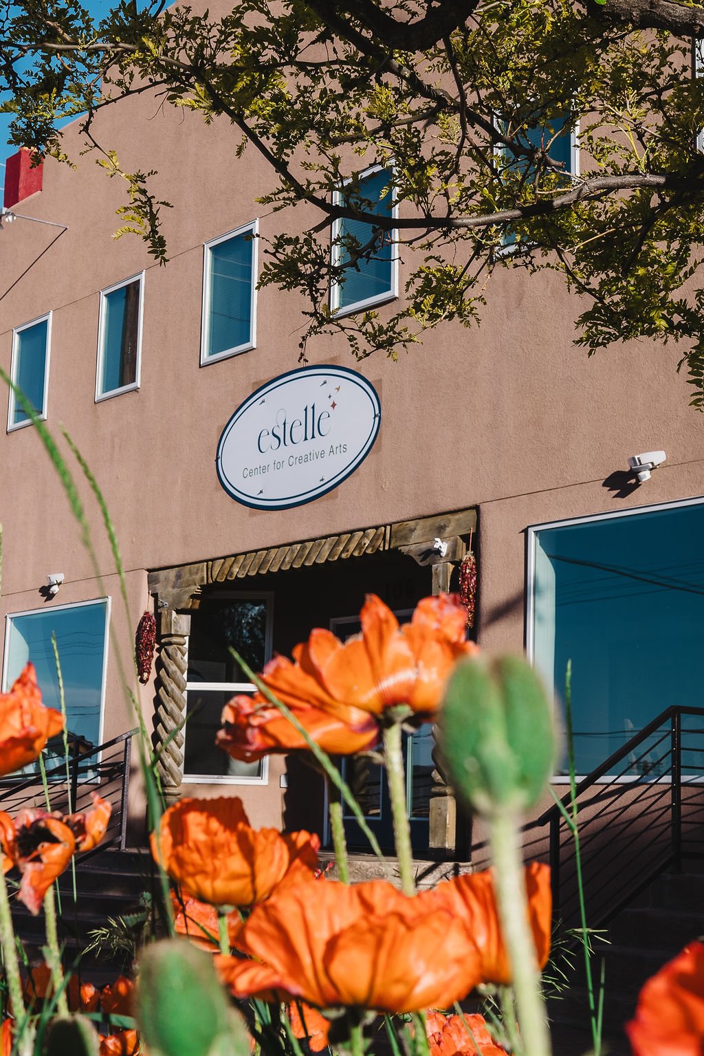 The photo shows the front of a building with a sign that reads 'estelle Center for Creative Arts.' There are orange flowers, possibly poppies, in the foreground and green foliage from a tree above. The building has multiple windows, and there are steps with a black railing leading up to the entrance.