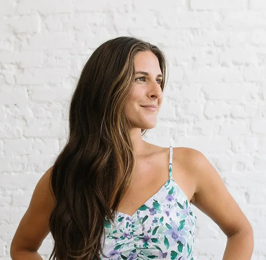 A woman with long, wavy brown hair is looking to her right, wearing a sleeveless floral dress with blue, purple, and green colors, standing against a white brick wall.