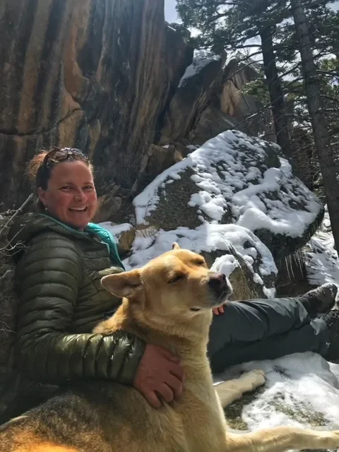 A woman smiling and sitting on snow-covered ground next to a dog, with a rocky, snow-dusted background and tall trees.